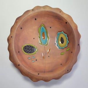 May include: A decorative wooden bowl with a scalloped edge. The bowl features three stylized flower designs in shades of teal, yellow, and black, with orange and green accents. Small black dots are scattered across the surface.
