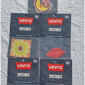 May include: Collection of Levi's iron-on patches in clear packaging. The patches feature various designs, including a crescent moon, a sunflower, red lips, a double-faced figure, and a postage stamp with a peace sign and the word "FREE".