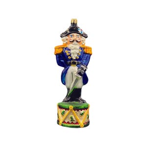 May include: A blue and gold glass Christmas ornament depicting a nutcracker soldier holding a sword and standing on a drum. The nutcracker has a white beard and mustache and is wearing a blue uniform with gold trim.