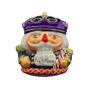 May include: A purple and gold Christmas ornament depicting a nutcracker holding a candy cane. The nutcracker has a white beard and mustache, and is wearing a black and gold uniform.