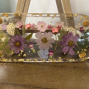May include: A clear resin tray with an oval shape, embedded with dried flowers in shades of purple, pink, and white. Gold leaf flakes are scattered throughout the resin, and around the edges. Decorative tray.