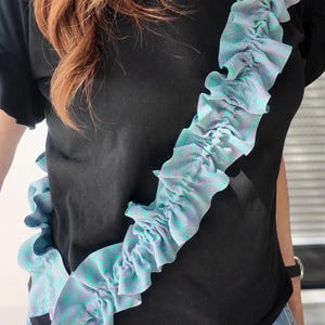 Peut inclure: Une bandoulière de téléphone en tissu à volants avec un motif marbré violet clair et bleu sarcelle. La sangle est fixée à une coque de téléphone transparente avec des clips en métal argenté, portée en diagonale sur un t-shirt noir et un jean bleu.