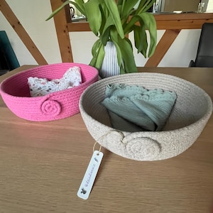 Può includere: Due cesti di corda fatti a mano in rosa e beige su un tavolo di legno. Il cesto rosa contiene un articolo in pizzo bianco, mentre il cesto beige contiene un panno verde piegato. Un'etichetta con testo è appesa al cesto beige.