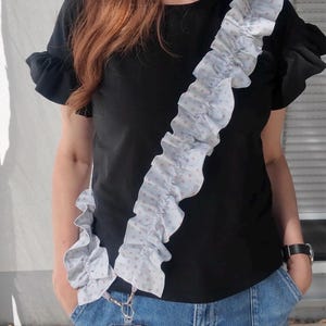 Könnte beinhalten: Ein hellblaues Handyband mit einem gerüschten Stoffdesign und einem dezenten rosa Blumenmuster. Das Band ist mit einem silberfarbenen Metallkarabiner an einer transparenten Schutzhülle befestigt und wird diagonal über einem schwarzen Oberteil getragen.