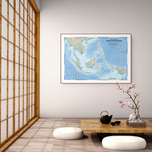 Puede incluir: Un mapa físico enmarcado titulado Island Southeast Asia que muestra la topografía detallada de la región en tonos verdes y azules, exhibido en una pared blanca sobre una mesa baja de madera con una tetera negra y una pequeña rama floreciente.
