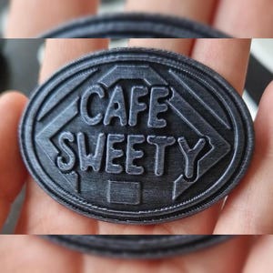 May include: A dark grey, oval emblem featuring the words "CAFE SWEETY" in raised lettering. The text is within a diamond shape, and the emblem has a textured surface.