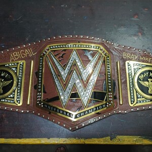 Może przedstawiać: Brązowo-złoty pas mistrzowski z logo WWE pośrodku. Pas zawiera napisy "ABIGAIL" i "BRAY WYATT" na bocznych płytach oraz "UNIVERSAL CHAMPION" na dole. Pas jest ozdobiony kryształkami.