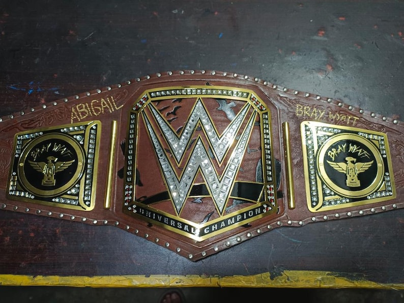 Może przedstawiać: Brązowo-złoty pas mistrzowski z logo WWE pośrodku. Pas zawiera napisy "ABIGAIL" i "BRAY WYATT" na bocznych płytach oraz "UNIVERSAL CHAMPION" na dole. Pas jest ozdobiony kryształkami.
