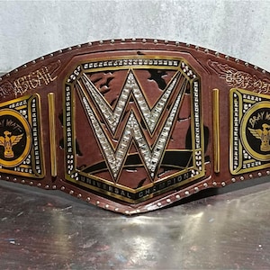 May include: A brown leather WWE Universal Championship belt with gold accents and the text "BRAY WYATT". The center features the WWE logo with embedded crystals. The belt is studded with rivets.