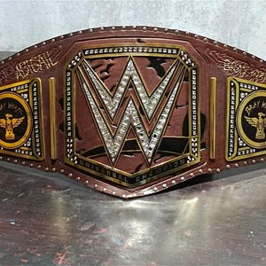 Może przedstawiać: Brązowy sk&oacute;rzany pas mistrzowski WWE Universal Championship. Centralna płyta przedstawia logo WWE z osadzonymi kryształkami. Boczne płyty wyświetlają "Bray Wyatt" i skrzydlaty emblemat. Pas jest wykończony złotymi i srebrnymi akcentami.