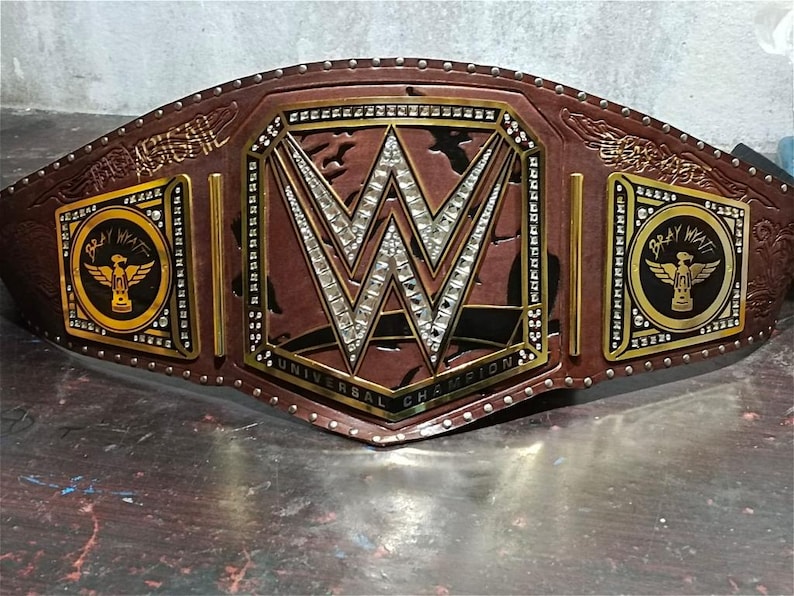 Może przedstawiać: Brązowy sk&oacute;rzany pas mistrzowski WWE Universal Championship. Centralna płyta przedstawia logo WWE z osadzonymi kryształkami. Boczne płyty wyświetlają "Bray Wyatt" i skrzydlaty emblemat. Pas jest wykończony złotymi i srebrnymi akcentami.