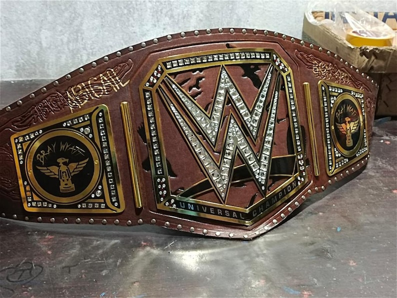 Może przedstawiać: Brązowy sk&oacute;rzany pas mistrzowski WWE Universal Championship ze złotymi i srebrnymi akcentami. Centralna płyta przedstawia logo WWE z osadzonymi kryształami. Pas zawiera tekst "UNIVERSAL CHAMPION" i "BRAY WYATT".