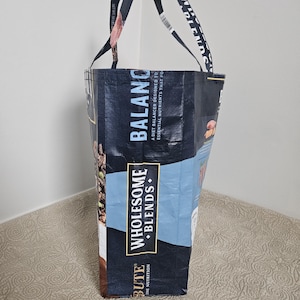 May include: A reusable tote bag made from a dark blue material with the words "BALANCE" and "WHOLESOME BLENDS" printed on it. The bag has black handles and is designed for carrying groceries or other items. The bag is made from a durable, recycled material.