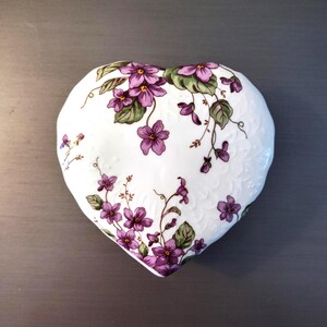 May include: A heart-shaped trinket dish with a white base, adorned with purple floral and green leaf patterns. The dish has a textured surface and is likely made of ceramic or porcelain.