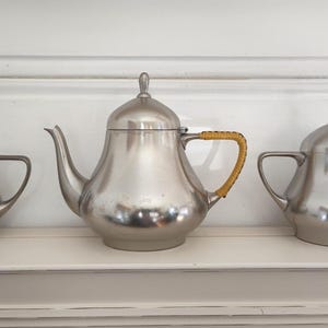 Puede incluir: Un juego de té plateado, que incluye una tetera con asa trenzada, una jarra para la crema y un azucarero. El juego se exhibe sobre una estantería blanca sobre un fondo blanco. La tetera tiene un pico curvo y una tapa con un pequeño pomo.