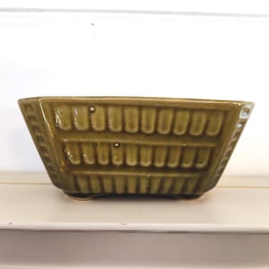 May include: A rectangular, olive-green ceramic planter with a textured, basket-weave design. The planter has a glossy finish and sits on four small feet. Ideal for small plants or succulents.