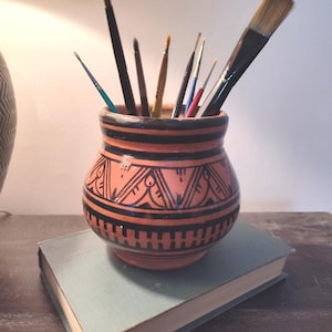 May include: A terracotta-colored ceramic pot with black geometric designs holds various paintbrushes. The pot sits atop a closed, light blue book. The paintbrushes are of different sizes and shapes, with wooden handles and bristles.