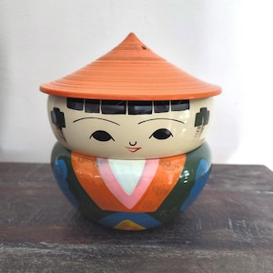 Può includere: Una bambola Kokeshi giapponese decorativa con un viso dipinto e un cappello conico arancione. Il corpo della bambola è dipinto con motivi geometrici colorati, tra cui arancione, blu e verde. La bambola è su una superficie di legno scuro.