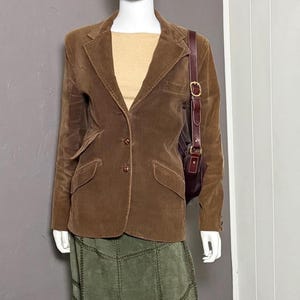 Puede incluir: Un maniquí exhibe una blazer de pana marrón, una blusa color beige y una falda de ante verde oliva. Un bolso de hombro burdeos completa el conjunto. La falda presenta costuras decorativas.