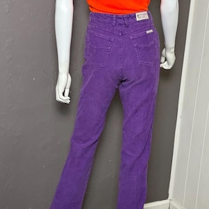 May include: A mannequin is wearing an orange vest top and purple corduroy trousers. The trousers have a high waist and a flared leg. The mannequin is standing on a black base against a grey wall.