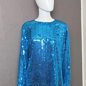 Peut inclure: Un haut turquoise à paillettes à manches longues. Le vêtement est présenté sur un mannequin blanc sur un fond gris. Les paillettes créent un effet scintillant, reflétant la lumière. Le haut a une encolure ronde et une coupe ample.