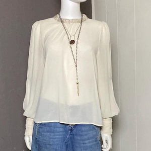 May include: A mannequin displays a cream-colored blouse with long sleeves and a denim skirt. The blouse has a beaded neckline and the skirt has a frayed hem. A layered necklace with a pendant is also visible.