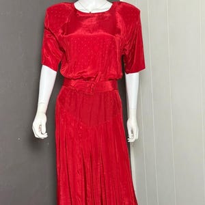 May include: A vibrant red dress on a mannequin. The dress features short sleeves, a belted waist, and a pleated skirt. The fabric has a subtle polka dot pattern. The dress is displayed on a white mannequin against a gray and white background.