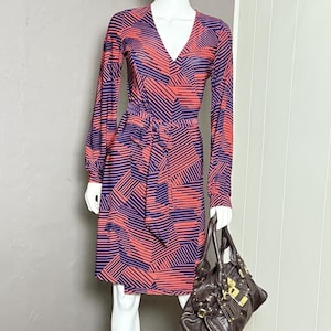 May include: A vintage-style dress with a V-neck and long sleeves, featuring a geometric pattern in red and purple. The dress is cinched at the waist with a matching belt. A brown leather handbag with gold hardware completes the look.