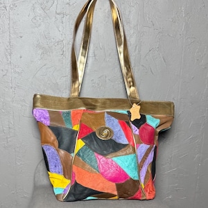 Könnte beinhalten: Eine bunte Patchwork-Leder-Tragetasche mit goldfarbenen Henkeln. Die Tasche hat Lederflicken in Lila-, Rot-, Orange-, Gelb- und Schwarztönen. Auf der Vorderseite befindet sich ein dekorativer Metallverschluss.