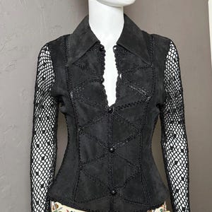 May include: A black long-sleeved shirt with a suede-like bodice featuring geometric stitched paneling and a button-down front. The sleeves are made of a black crocheted mesh material. The shirt is paired with blue denim jeans and a cream belt with a colorful floral pattern.