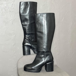 May include: A pair of black knee-high boots with a platform sole and block heels. The boots are made of a shiny, smooth material and are displayed on a light gray chair against a textured gray background.