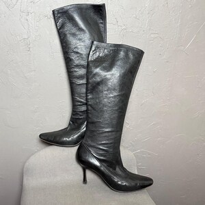 May include: A pair of black knee-high boots featuring a textured leather finish, a pointed toe, and a slim stiletto heel. The boots have a tall, structured shaft that reaches the knee, with a smooth, fitted silhouette and a sleek, polished appearance.