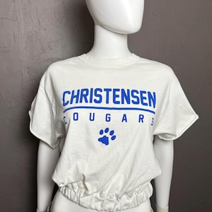 May include: A white short-sleeved t-shirt with a gathered elastic hem. The front features the blue text CHRISTENSEN COUGARS and a blue paw print graphic. The shirt is displayed on a mannequin wearing blue jeans with a patterned belt.