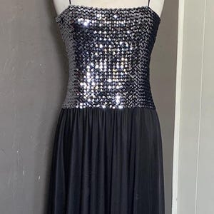 Peut inclure: Robe de soirée noire, longue, sur un mannequin. Le corsage est recouvert de paillettes argentées et la jupe est en tissu noir fluide. La robe a de fines bretelles et un pendentif en forme de cœur.