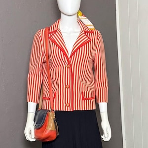 May include: A mannequin wearing a vintage ensemble. The outfit features a red and white striped jacket, a black pleated skirt, and a multi-coloured handbag. A patterned scarf is worn on the head. The jacket has gold buttons and red trim.