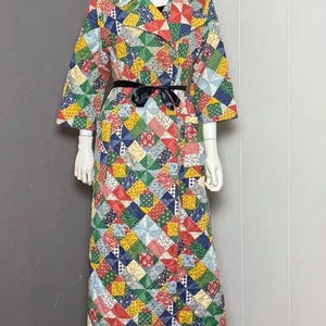 May include: A long, quilted coat featuring a vibrant patchwork pattern of small squares in red, yellow, blue, green, and white. The coat has a wide collar, three-quarter length sleeves, and a black ribbon tie at the waist over a black skirt.