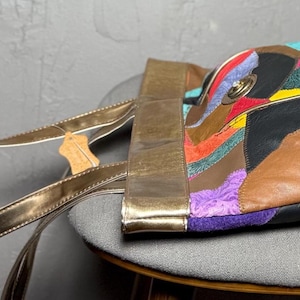 Peut inclure: Un sac fourre-tout en cuir patchwork multicolore avec des anses dor&eacute;es. Le sac pr&eacute;sente des pi&egrave;ces de cuir de couleurs rouge, violet, jaune, marron et noir. Le sac est pos&eacute; sur un tabouret gris.