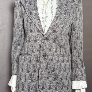 May include: A gray and brown patterned blazer with a two-button closure and notched lapels, worn over a white collared shirt featuring a ruffled front placket and ruffled cuffs that extend past the sleeves of the blazer.