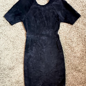May include: A black suede dress with short sleeves and a scoop neckline. The dress has a fitted silhouette and two pockets. The fabric has a textured appearance. The dress is suitable for various occasions.
