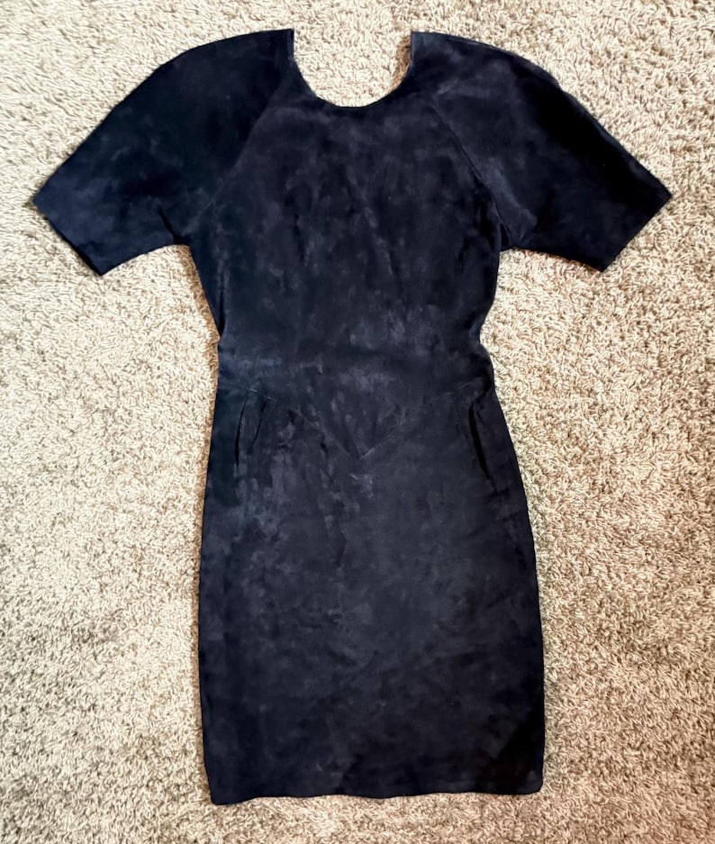 May include: A black suede dress with short sleeves and a scoop neckline. The dress has a fitted silhouette and two pockets. The fabric has a textured appearance. The dress is suitable for various occasions.