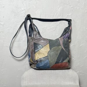 May include: A patchwork leather shoulder bag in various colors, including blue, red, and tan. The bag has a black strap and a curved shape, resting on a neutral-colored surface.