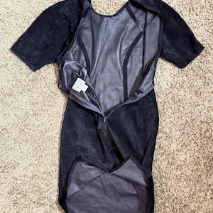 May include: A black suede dress with short sleeves and a knee-length hem. The dress has a zipper down the front and a scoop neckline. The interior lining is a lighter shade of gray. The dress is laid flat on a neutral-colored carpet.