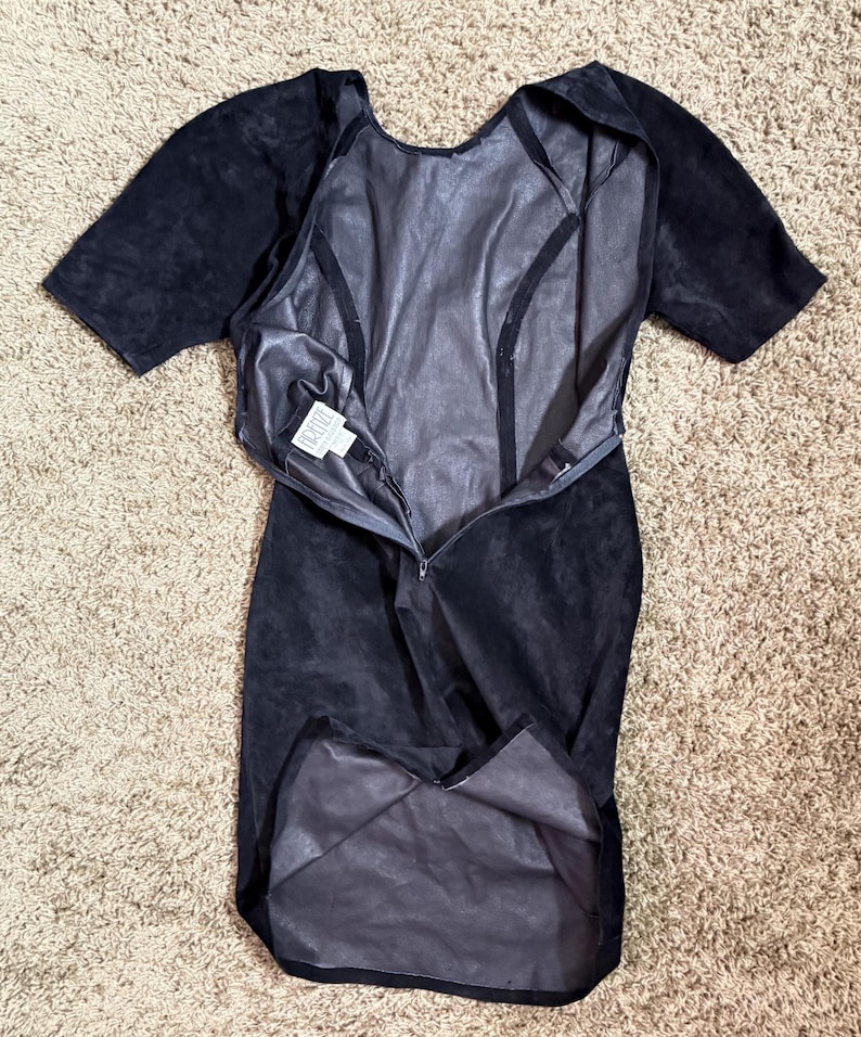 May include: A black suede dress with short sleeves and a knee-length hem. The dress has a zipper down the front and a scoop neckline. The interior lining is a lighter shade of gray. The dress is laid flat on a neutral-colored carpet.