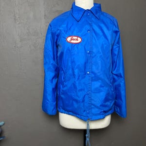 Op de afbeelding: Een blauwe nylon jas met een drukknoopsluiting en een trekkoord aan de onderkant. De jas heeft een patch op de linkerborst met de tekst "Jack" in rode letters.