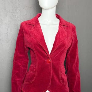 May include: A vibrant red velvet blazer featuring a notched lapel, long sleeves, and a single red button closure at the center. The jacket includes two front pockets with flaps and visible stitching along the seams, tailored for a fitted silhouette.
