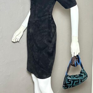 May include: A black suede dress with short sleeves and a wrap-style skirt is displayed on a mannequin. The mannequin also holds a small, sequined handbag with a geometric pattern in shades of teal and black.