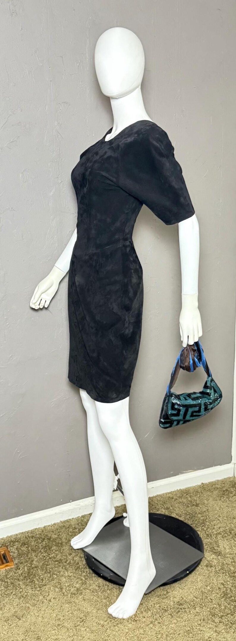 May include: A black suede dress with short sleeves and a wrap-style skirt is displayed on a mannequin. The mannequin also holds a small, sequined handbag with a geometric pattern in shades of teal and black.