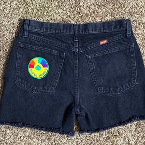 Pode incluir: Shorts de ganga preta com bainha desfiada e um remendo arco-íris que diz "Super Troop 9" no bolso de trás.