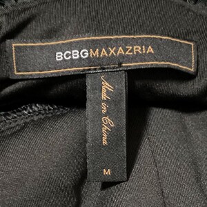 K&ouml;nnte beinhalten: Nahaufnahme eines schwarzen Kleidungsst&uuml;cketiketts mit dem Markennamen "BCBGMAXAZRIA" in goldfarbenen Buchstaben auf einem braunen rechteckigen Etikett. Darunter befindet sich ein kleineres schwarzes Etikett mit dem Text "Made in China" und der Gr&ouml;&szlig;e "M".