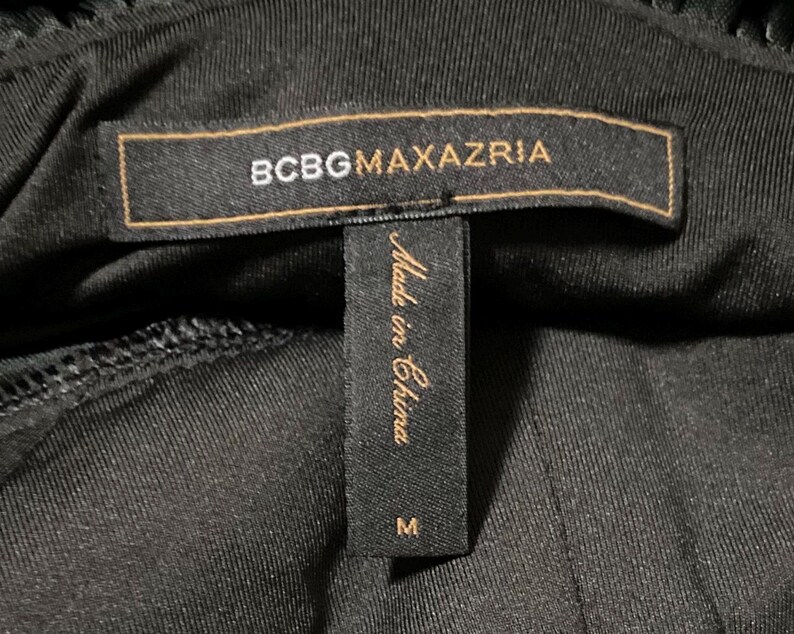 K&ouml;nnte beinhalten: Nahaufnahme eines schwarzen Kleidungsst&uuml;cketiketts mit dem Markennamen "BCBGMAXAZRIA" in goldfarbenen Buchstaben auf einem braunen rechteckigen Etikett. Darunter befindet sich ein kleineres schwarzes Etikett mit dem Text "Made in China" und der Gr&ouml;&szlig;e "M".